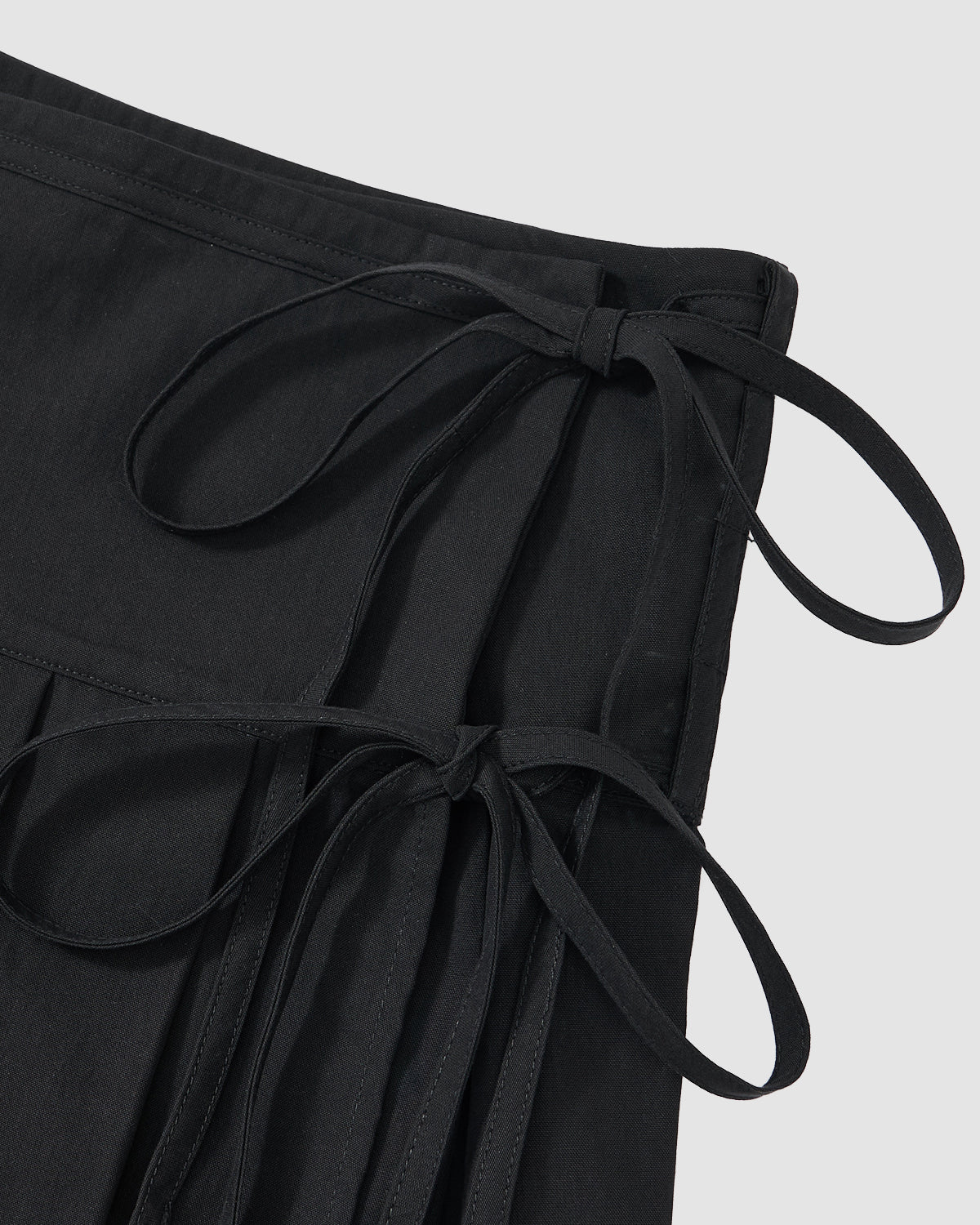 Close-up of the BLACK UNIFORM APRON SKIRT by Sundae School in black, with two fabric tie bows on the waistband and side, shown against a plain light gray background.