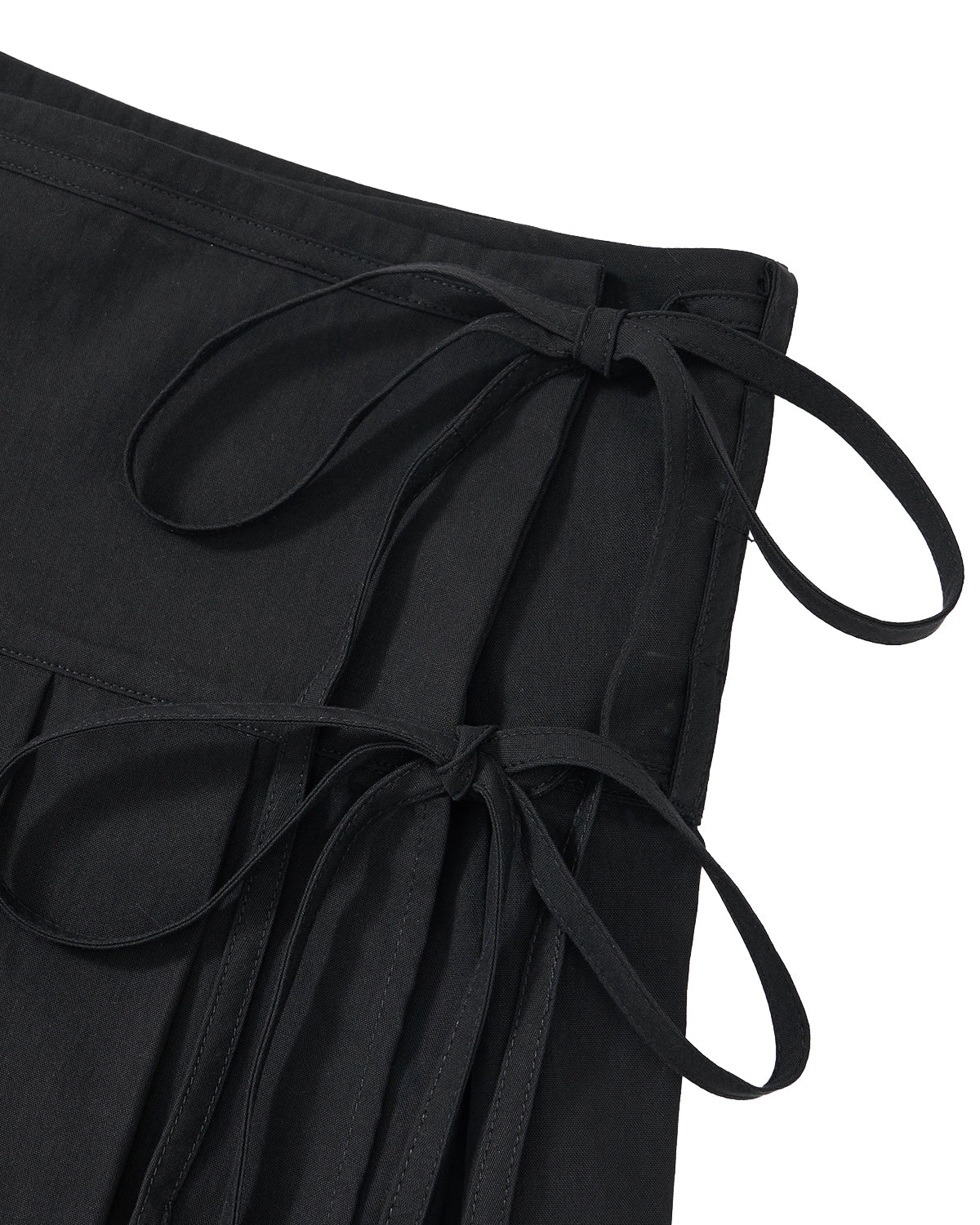 Close-up of the Sundae School BLACK UNIFORM APRON SKIRT, a black pleated skirt inspired by Korean-style aprons. It features two fabric ties on the waistband, neatly knotted into bows, and is crafted from cotton or a similar material with visible stitching along the edges.