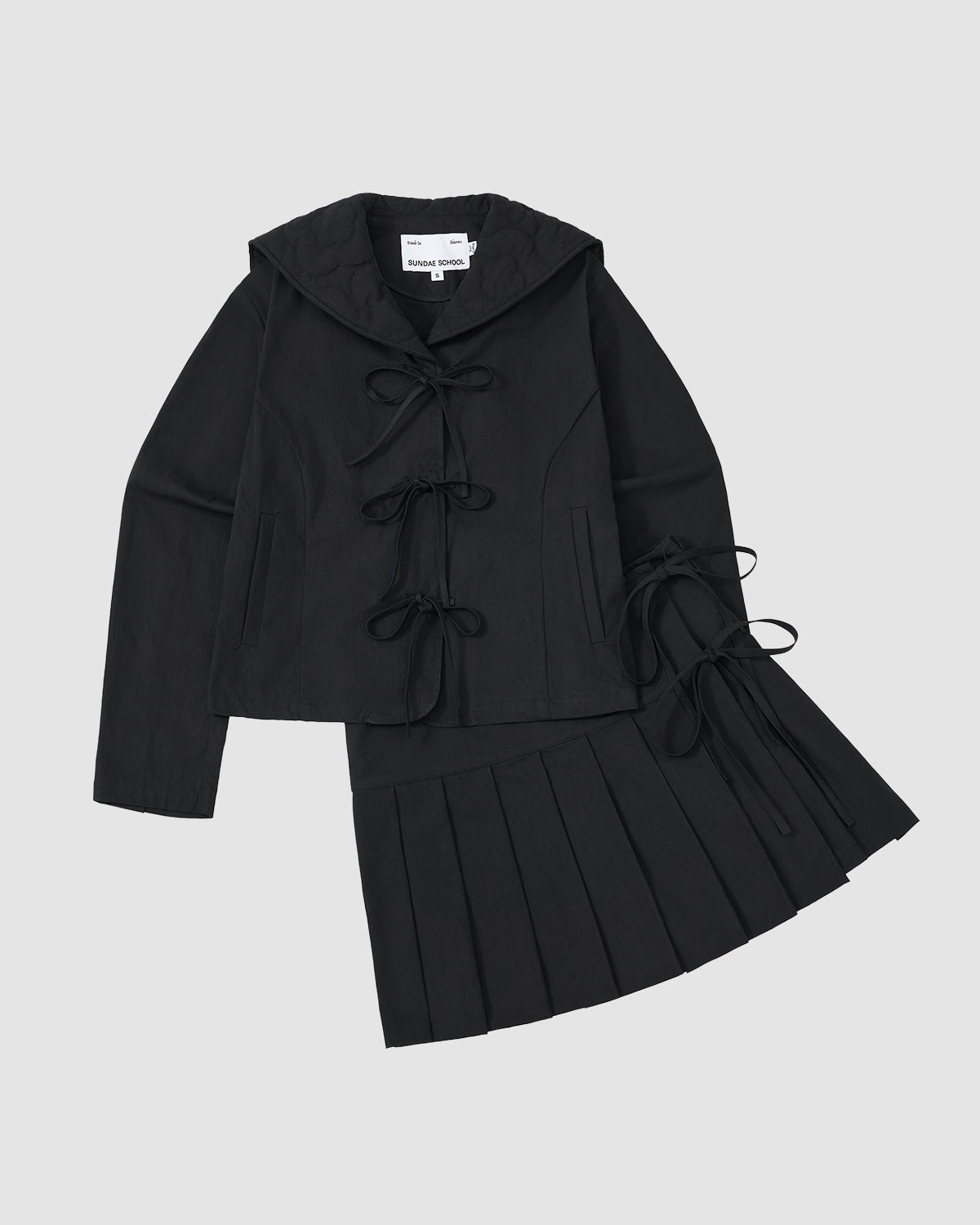 The Sundae School BLACK UNIFORM APRON SKIRT set features a black hooded jacket with three front bows and a pleated skirt, inspired by Korean apron styles, both displayed flat on a light gray background.