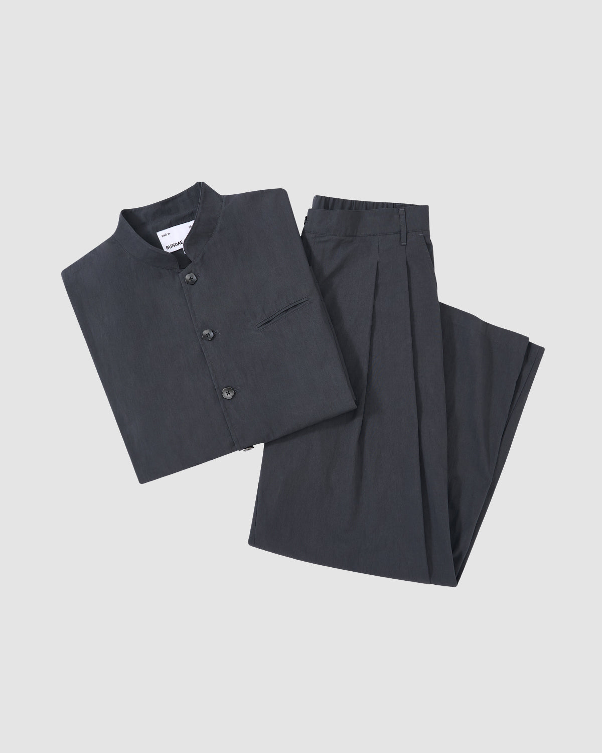 The CHARCOAL STAND COLLAR UNIFORM JACKET by Sundae School, made from a cotton nylon blend with buttons and a chest pocket, is neatly displayed with matching pleated trousers on a light gray background.