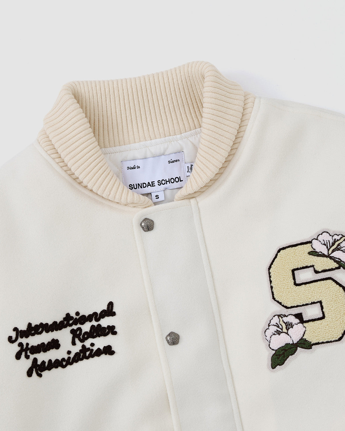 The Sundae School IVORY VARSITY JACKET features an oversized cream design with vegan leather sleeves, a ribbed collar, embroidered International Home Roller Association text, a yellow S patch with white flowers, and an inner label.