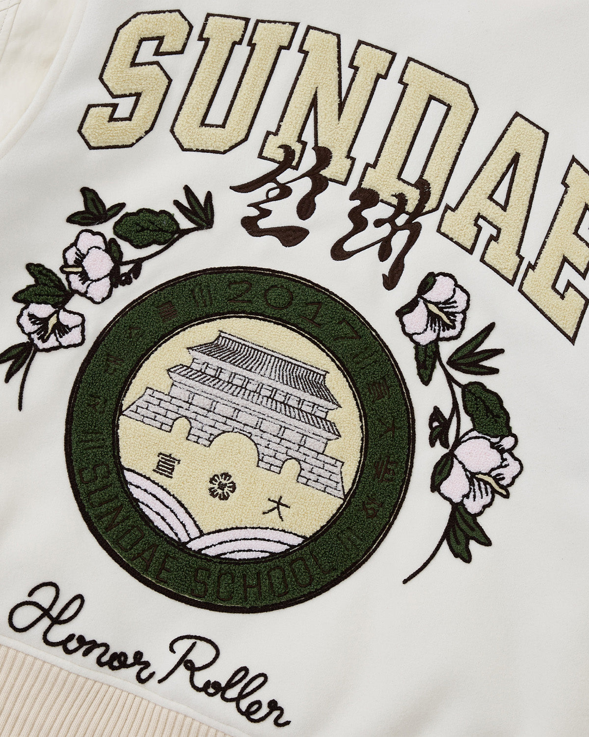 Close-up of the Sundae School IVORY VARSITY JACKET, an oversized design with white fabric, vegan leather sleeves, embroidered SUNDAE and Honor Roller text, plus a green emblem featuring an Asian-style building and floral motifs.