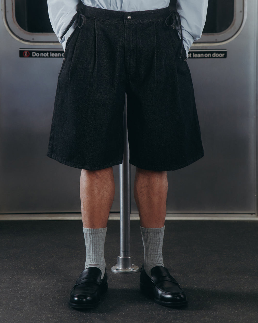 A person stands on a subway, wearing Sundae School BLACK VITA JORTS, light gray socks, and black loafers. Only their lower half is visible against a backdrop of a metal subway door with a warning sign.
