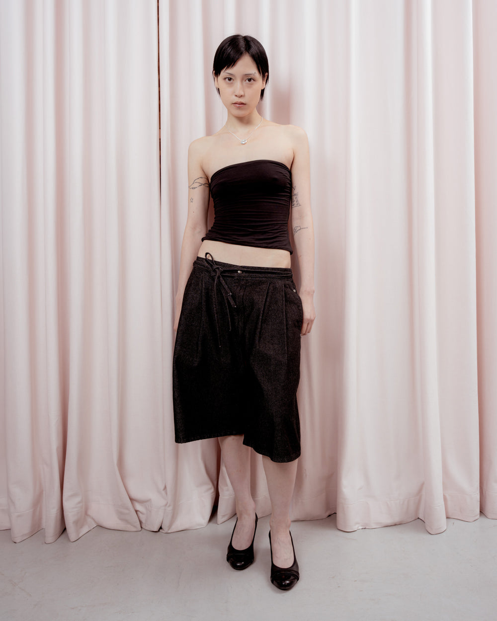 A person with short dark hair stands in front of pale pink curtains, wearing a strapless black top, the BLACK VITA JORTS by Sundae School—knee-length pleated denim jorts with a wide-leg silhouette—black heels, and a necklace. Their arms are relaxed by their sides.
