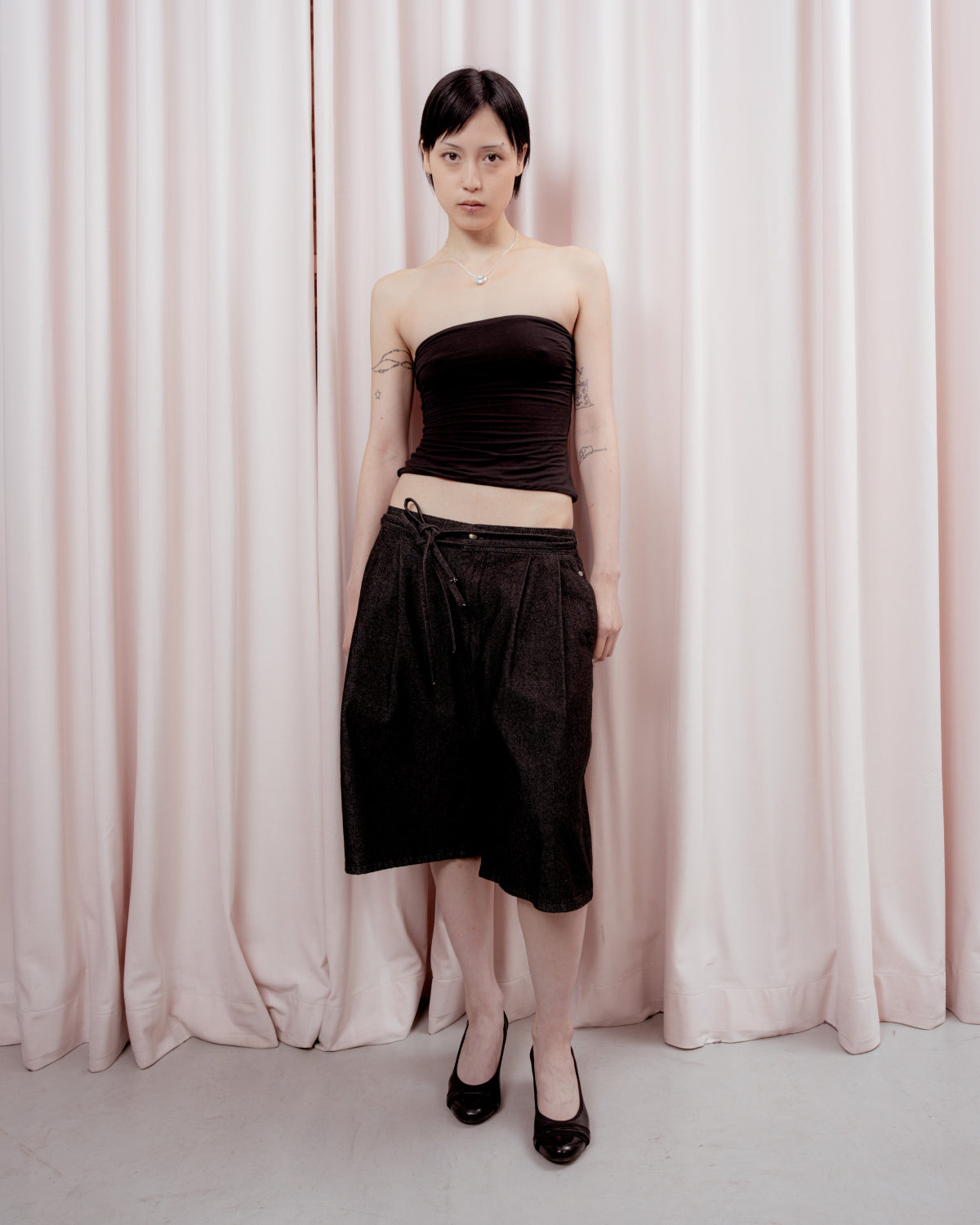 A person with short dark hair stands in front of pale pink curtains, wearing a strapless black top, the BLACK VITA JORTS by Sundae School—knee-length pleated denim jorts with a wide-leg silhouette—black heels, and a necklace. Their arms are relaxed by their sides.