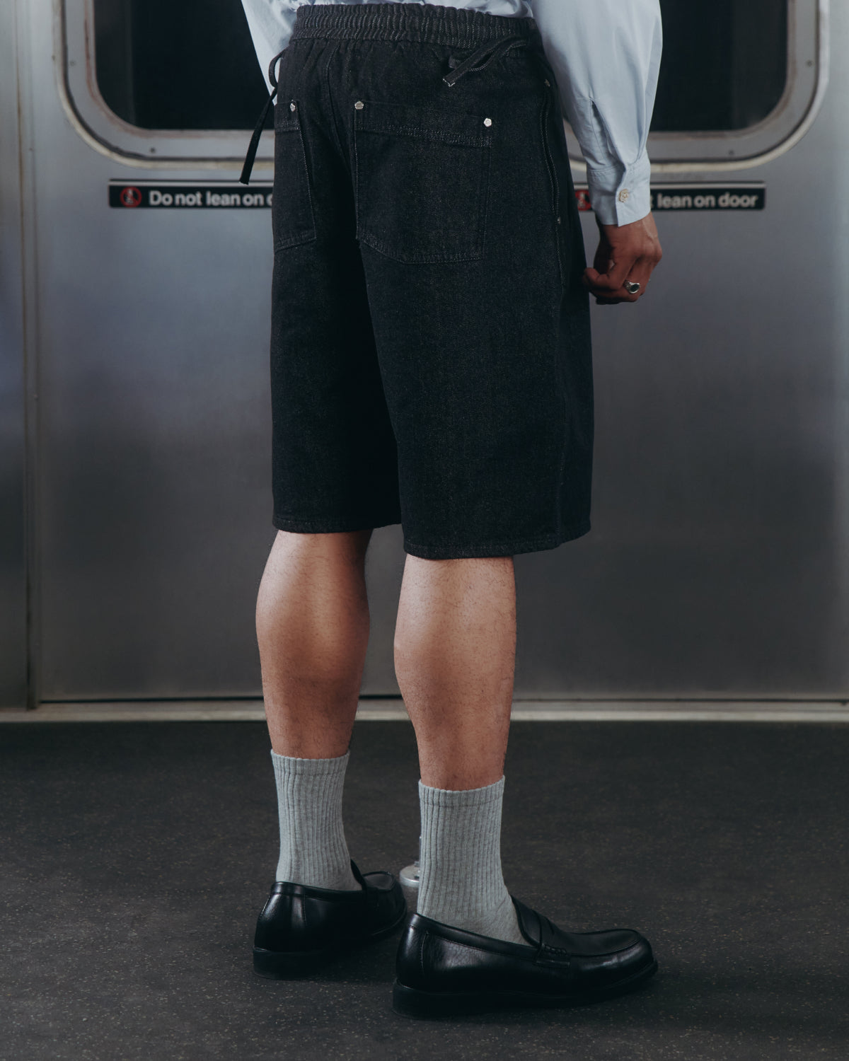A person in a light blue shirt, Sundae School BLACK VITA JORTS, gray socks, and black shoes stands facing away on a subway train with a 
