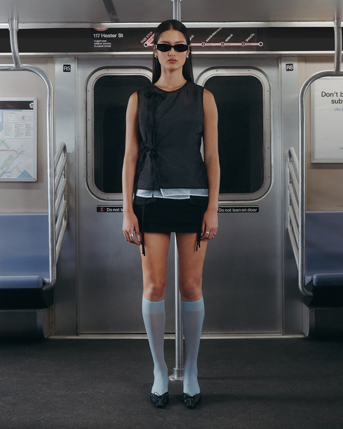 A woman wears Sundae School's BLACK VITA ORGANZA VEST, styled with a black mini skirt and light blue knee-high socks, standing in a subway car beneath the 117 Hester St. sign and facing the camera with a neutral expression.