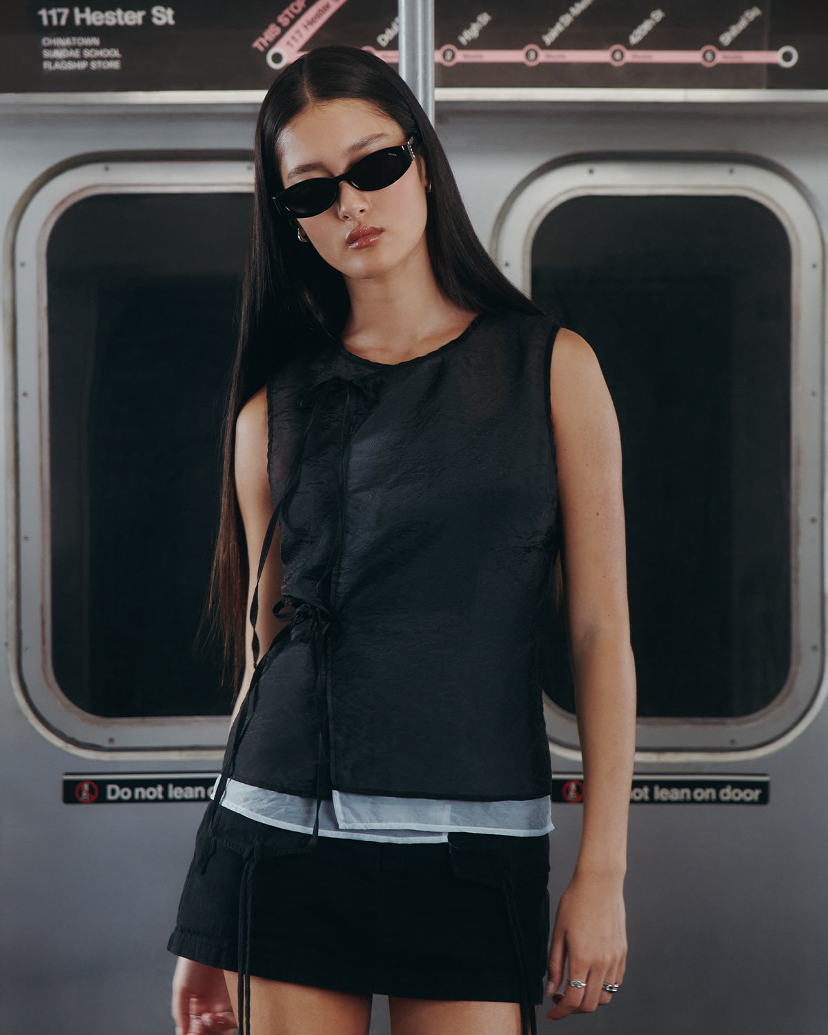 A woman with long dark hair and black sunglasses, wearing the Sundae School BLACK VITA ORGANZA VEST over sky blue with a black skirt, stands by subway doors marked 