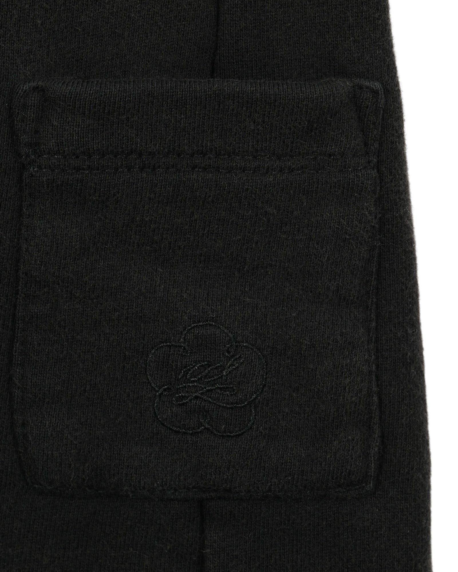 Close-up of the BLACK WASH WRAP SWEAT BAJI by Sundae School, showcasing a black fabric pocket with subtle embroidered flower and script details on its soft, high-quality cotton surface—reminiscent of unisex jogger sweatpants.