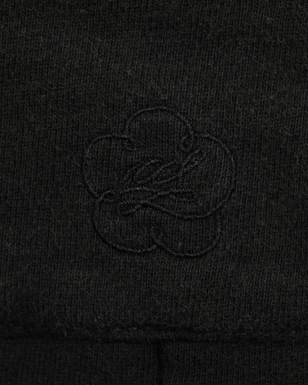 Close-up of the black fabric from the Sundae School BLACK WASH WRAP SWEAT BAJI, featuring a subtle, stitched clover-shaped logo design in the center. The texture of the material and the embroidery are clearly visible, highlighting the quality craftsmanship typical of these premium cotton sweatpants.