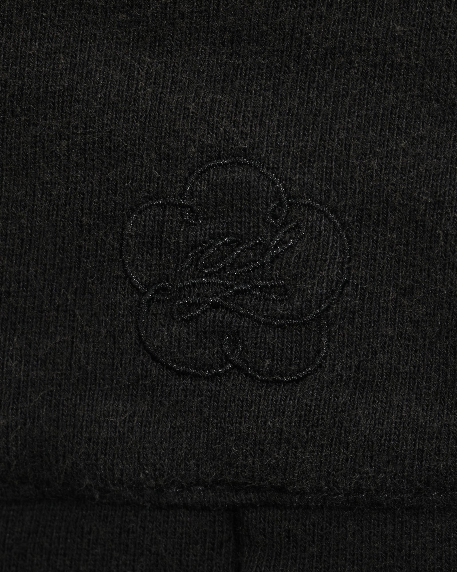 Close-up of the black fabric from the Sundae School BLACK WASH WRAP SWEAT BAJI, featuring a subtle, stitched clover-shaped logo design in the center. The texture of the material and the embroidery are clearly visible, highlighting the quality craftsmanship typical of these premium cotton sweatpants.