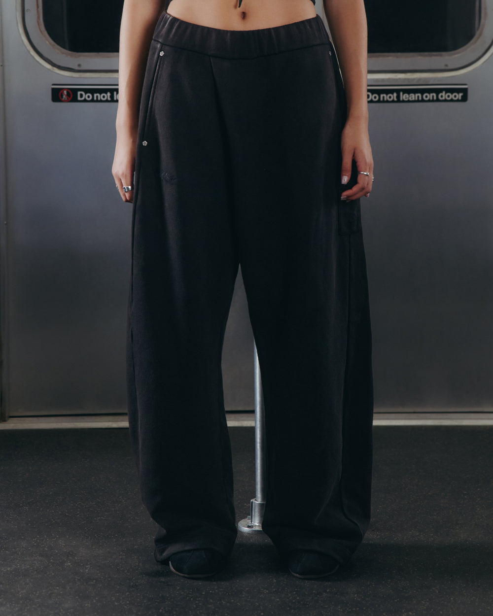 A person wearing Sundae School’s BLACK WASH WRAP SWEAT BAJI and black shoes stands inside a subway car with their hands at their sides. The upper body is not visible, and signs on the subway door behind read “Do not block” and “Do not lean on door.”.