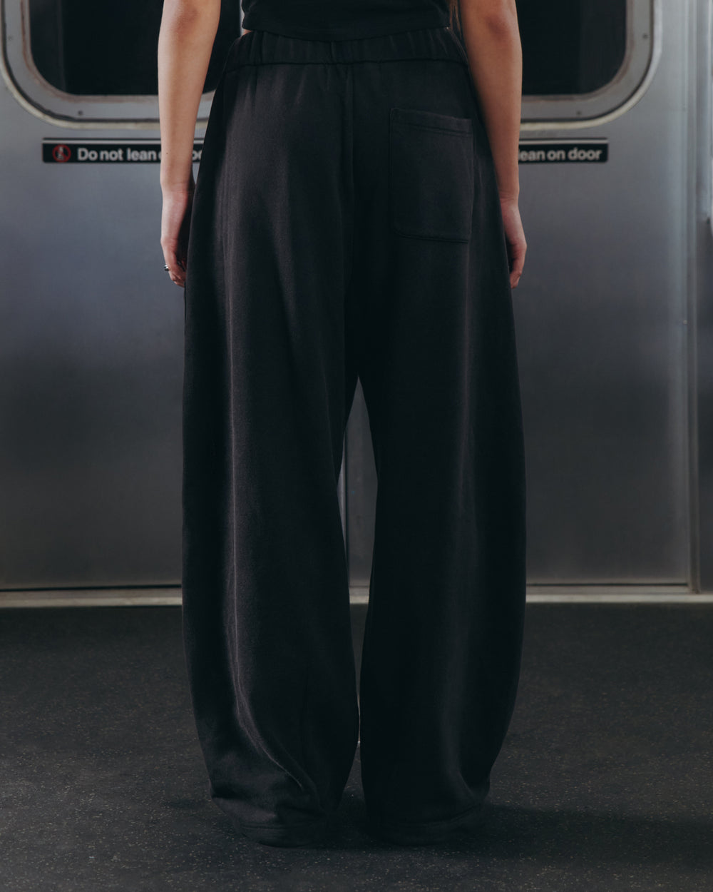 A person wearing Sundae School’s BLACK WASH WRAP SWEAT BAJI stands with their back to the camera on a subway train, facing closed metal doors adorned with warning signs.