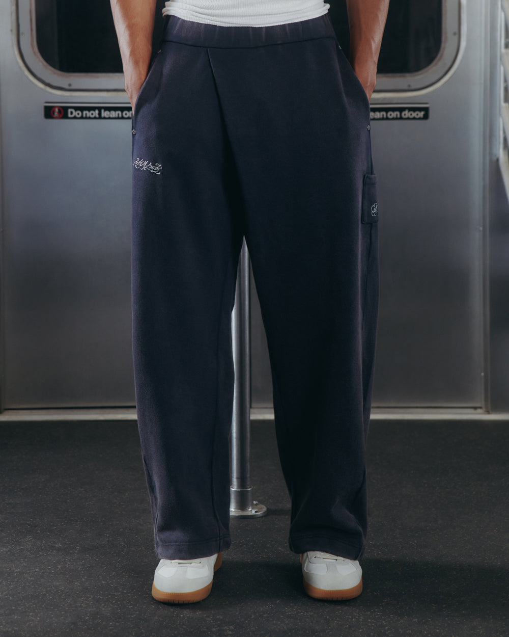 A person wearing Sundae School’s NAVY WASH WRAP SWEAT BAJI joggers and white sneakers stands inside a subway train. The loose, dark navy unisex joggers feature subtle embroidery, while the background includes part of a subway door with signage.