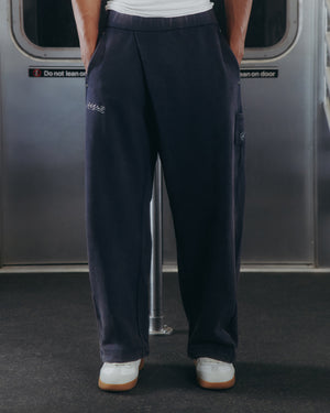 A person wearing Sundae School’s NAVY WASH WRAP SWEAT BAJI joggers and white sneakers stands inside a subway train. The loose, dark navy unisex joggers feature subtle embroidery, while the background includes part of a subway door with signage.