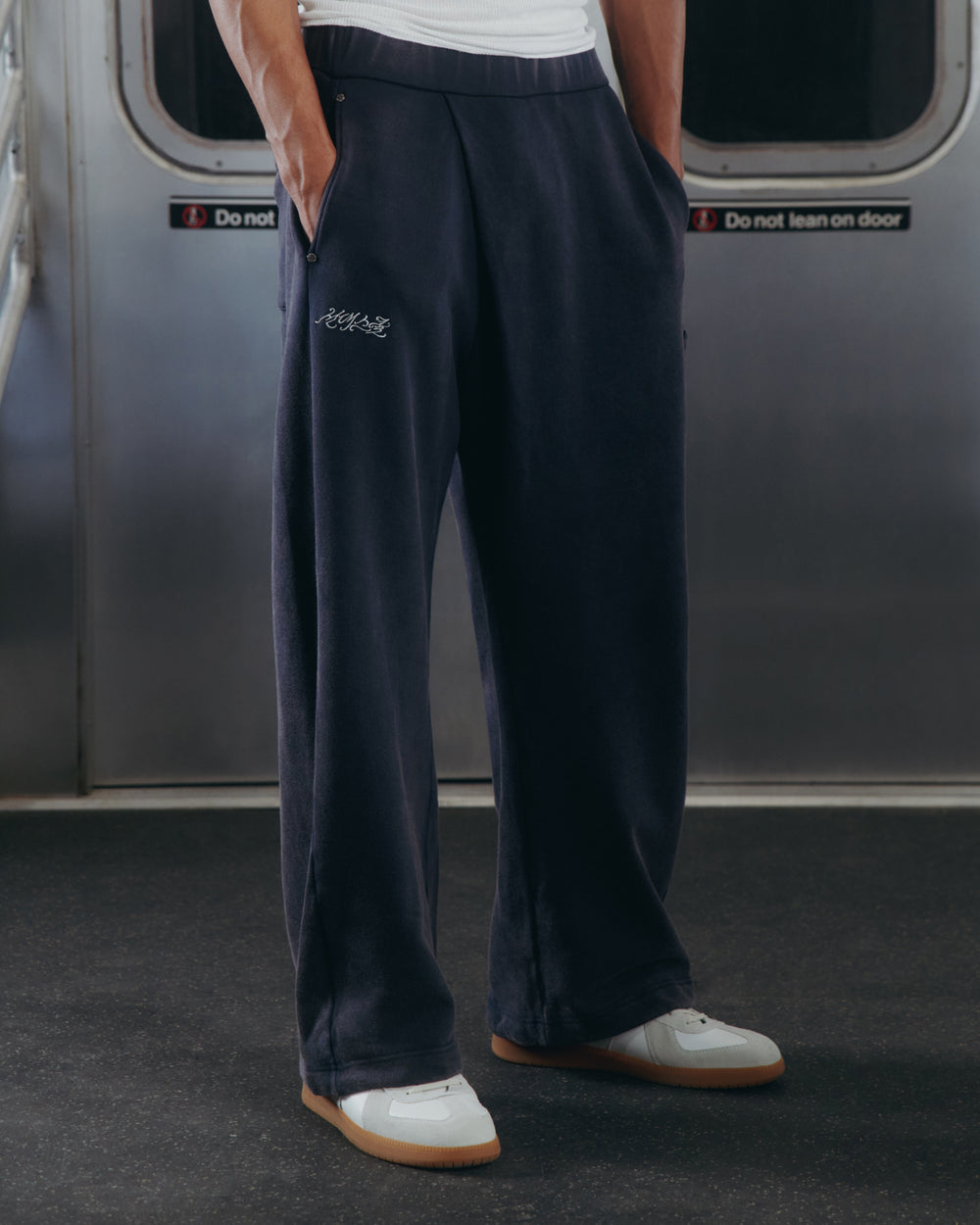 A person wearing Sundae School's NAVY WASH WRAP SWEAT BAJI with white sneakers stands on a subway train, hands in pockets, as "Do not lean on door" signs are visible behind them.