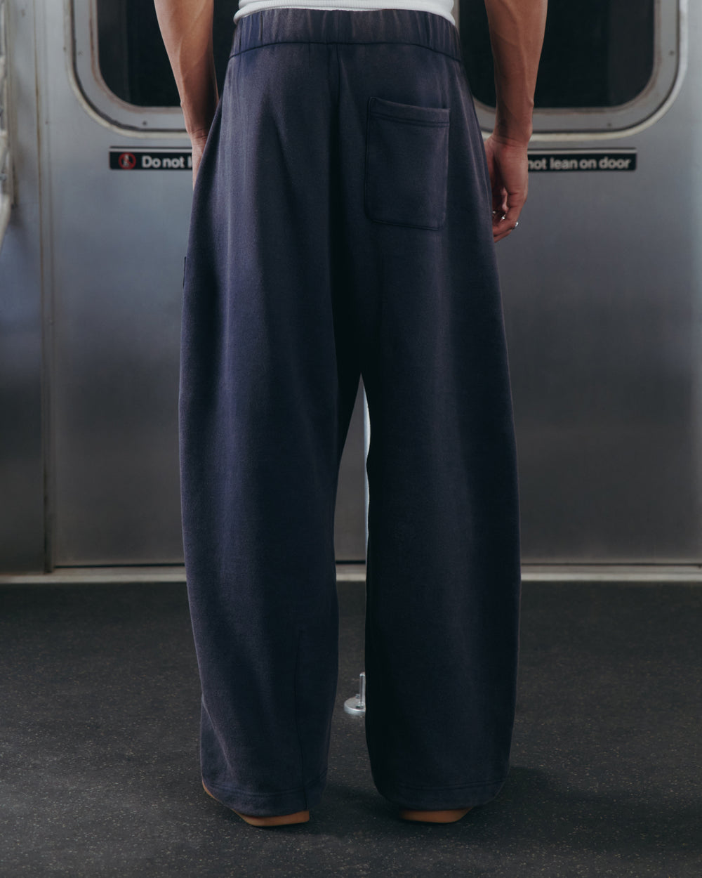 A person wearing Sundae School's NAVY WASH WRAP SWEAT BAJI stands with their back to the camera inside a subway train, where signs on the door read “Do not lean on door.”.