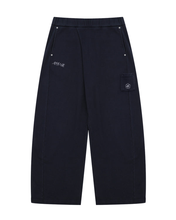 NAVY WASH WRAP SWEAT BAJI by Sundae School features a wide-leg unisex silhouette in black, an elastic waistband, two front pockets with button closures, subtle white embroidered text on one side, and a flap pocket adorned with a flower patch on the other.