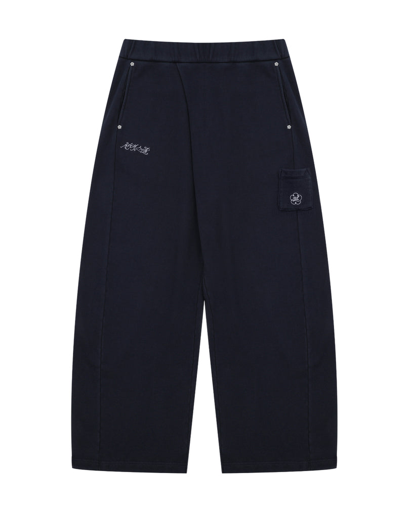 NAVY WASH WRAP SWEAT BAJI by Sundae School features a wide-leg unisex silhouette in black, an elastic waistband, two front pockets with button closures, subtle white embroidered text on one side, and a flap pocket adorned with a flower patch on the other.
