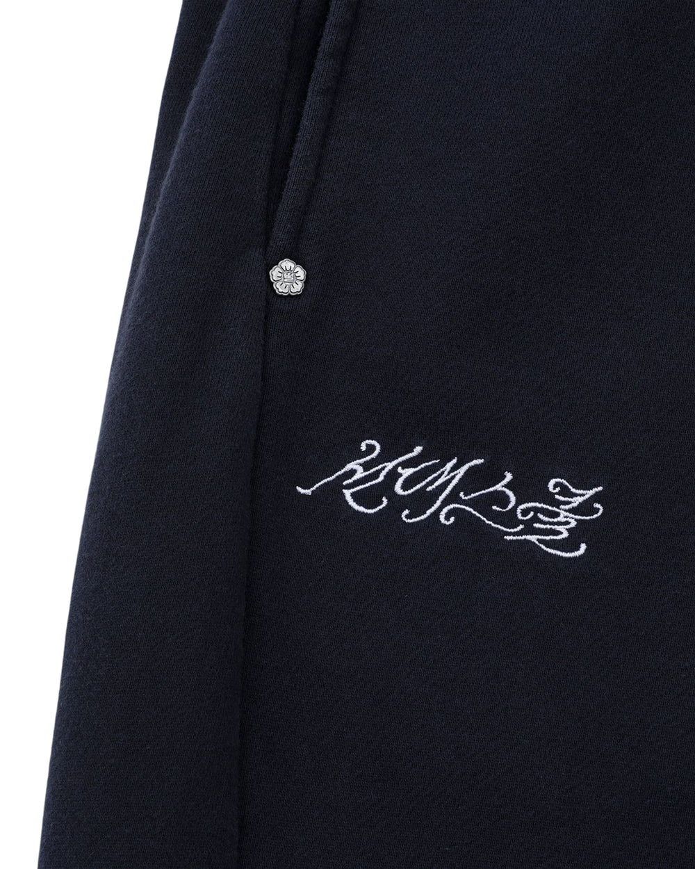 Close-up of Sundae School's NAVY WASH WRAP SWEAT BAJI––navy cotton sweatpants featuring a silver decorative button near the pocket and ornate white embroidery reading "HUMANZ" on the leg.
