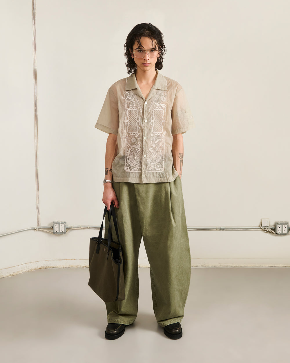 A person with short, wavy hair, glasses, and tattoos stands indoors against a neutral background. They wear a beige embroidered shirt and olive-green pants, holding Sundae School's OLIVE BOJAGI TIE TOTE BAG.