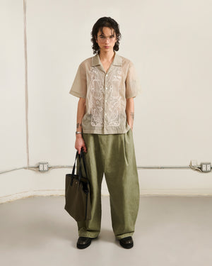 A person with short, wavy hair, glasses, and tattoos stands indoors against a neutral background. They wear a beige embroidered shirt and olive-green pants, holding Sundae School's OLIVE BOJAGI TIE TOTE BAG.