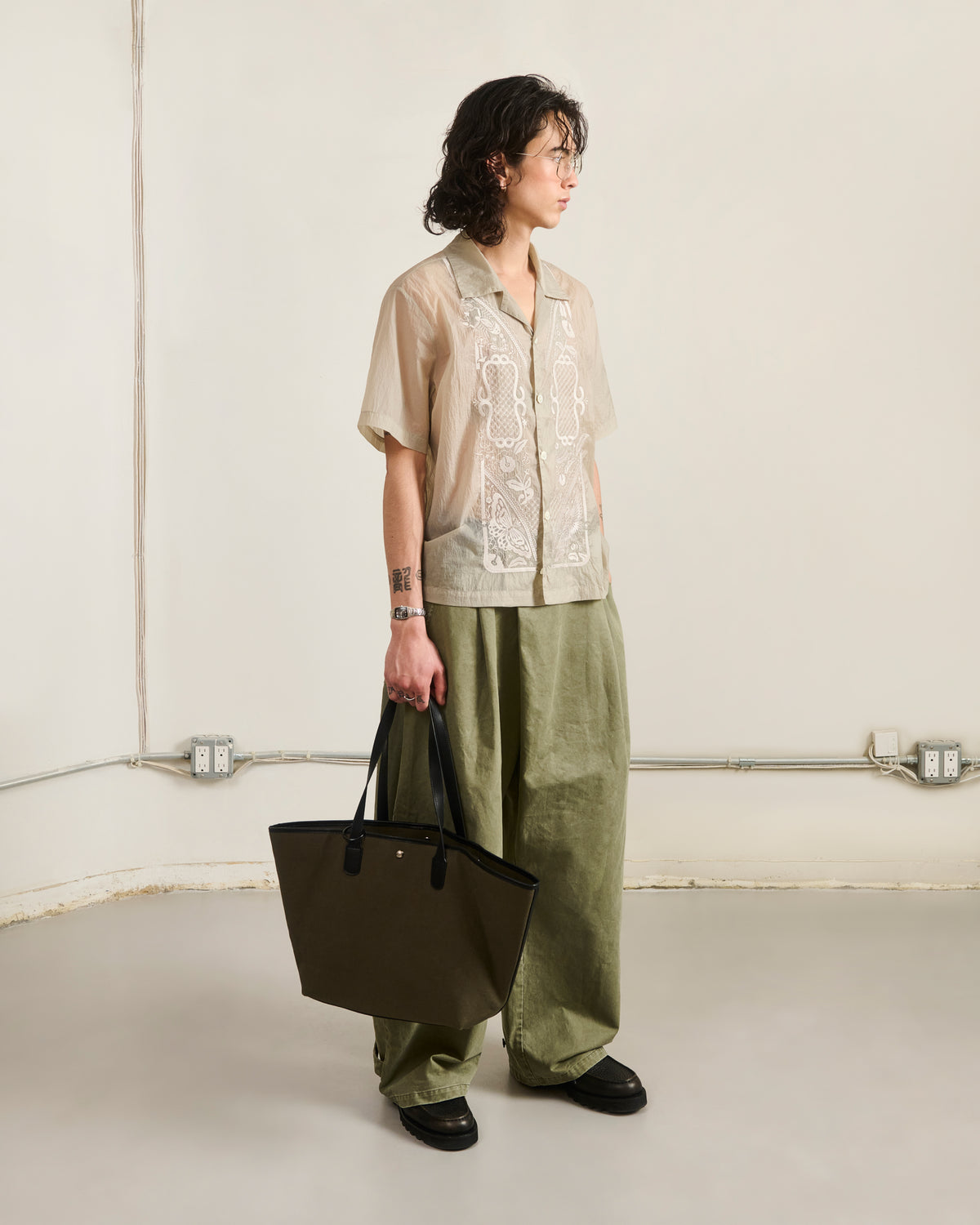 A person with wavy hair stands in a minimalist room, wearing a sheer embroidered shirt, loose olive green pants, black shoes, and holding the OLIVE BOJAGI TIE TOTE BAG by Sundae School.