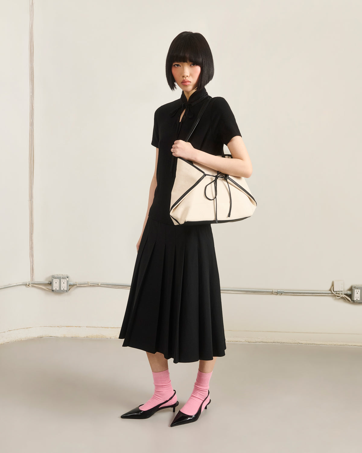 A woman with a short black bob wears a black dress, pink socks, and pointed black heels. She carries the Sundae School IVORY BOJAGI TIE TOTE BAG with vegan leather handles, standing against a plain wall with exposed outlets and pipes.
