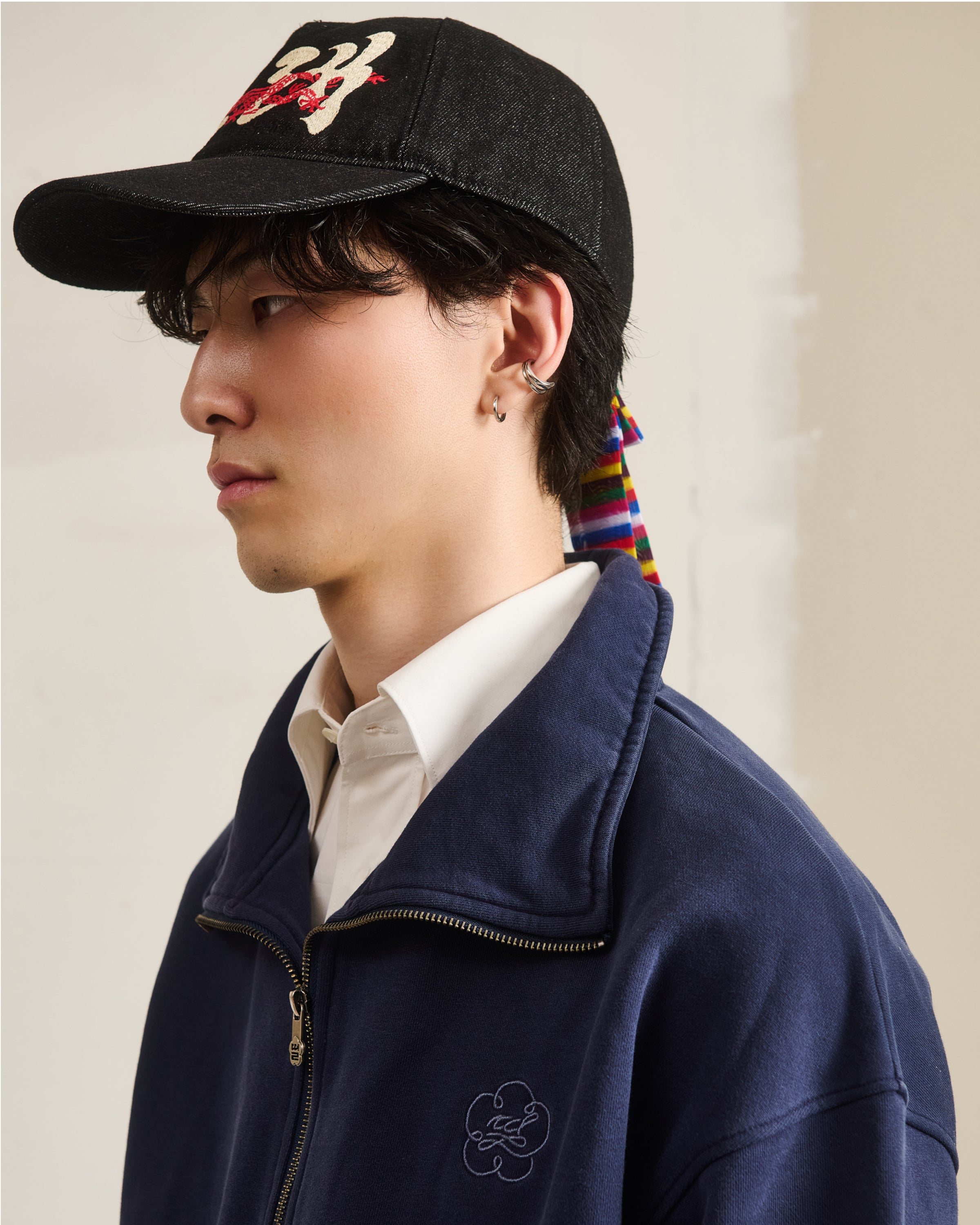 A young person in a dark cap with colorful ribbon, white collared shirt, and an oversized Sundae School NAVY HESTER QUARTER ZIP with clover logo stands in profile against a light background.