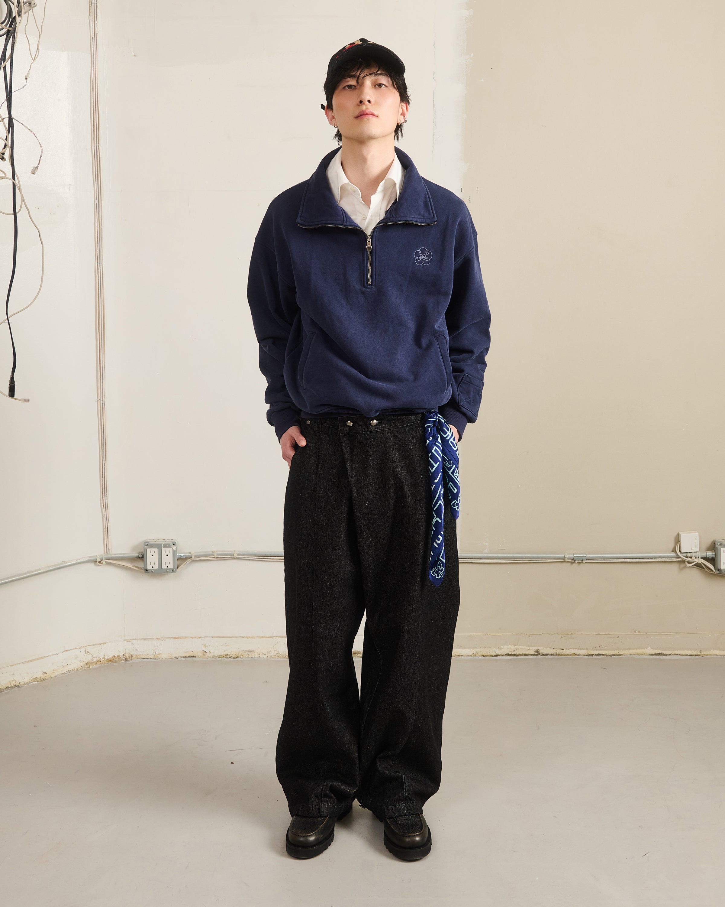 Indoors, a person wears a black cap, white collared shirt, Sundae School NAVY HESTER QUARTER ZIP sweatshirt, wide black pants with a blue bandana at the waist, and black shoes. The plain background shows wires and outlets.