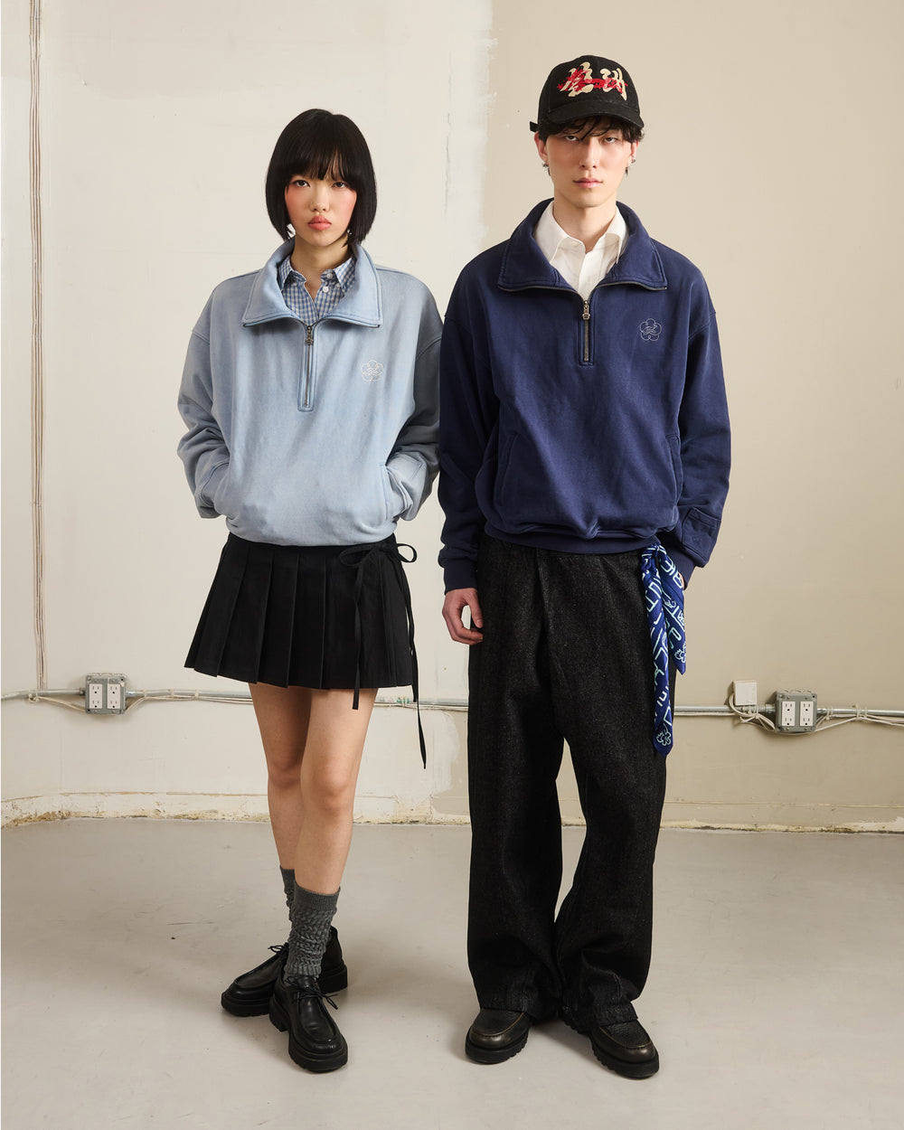 Two people stand side by side against a plain wall. The person on the left wears the SKY BLUE HESTER QUARTER ZIP by Sundae School, a black pleated skirt, gray socks, and black shoes. The person on the right sports a navy sweat jersey, dark wide pants, black shoes, and a black cap.