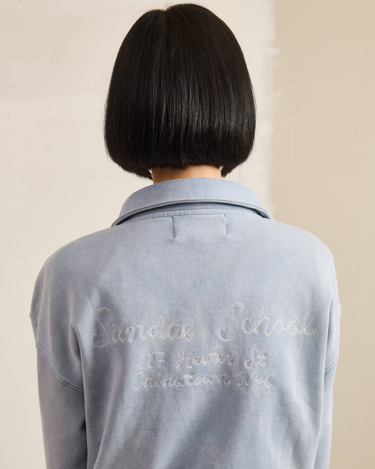 A person with a straight black bob is shown from behind, wearing the Sundae School SKY BLUE HESTER QUARTER ZIP, featuring cursive embroidery reading "Sundae School, 17 Baxter St, Chinatown, NYC.