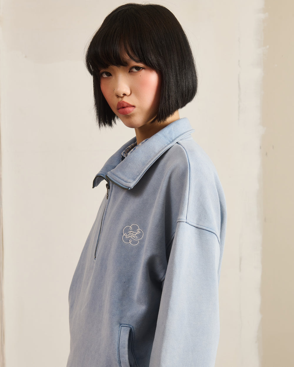 A person with a short black bob wears the Sundae School SKY BLUE HESTER QUARTER ZIP, featuring a small embroidered design, standing before a plain light background and looking at the camera with a neutral expression.