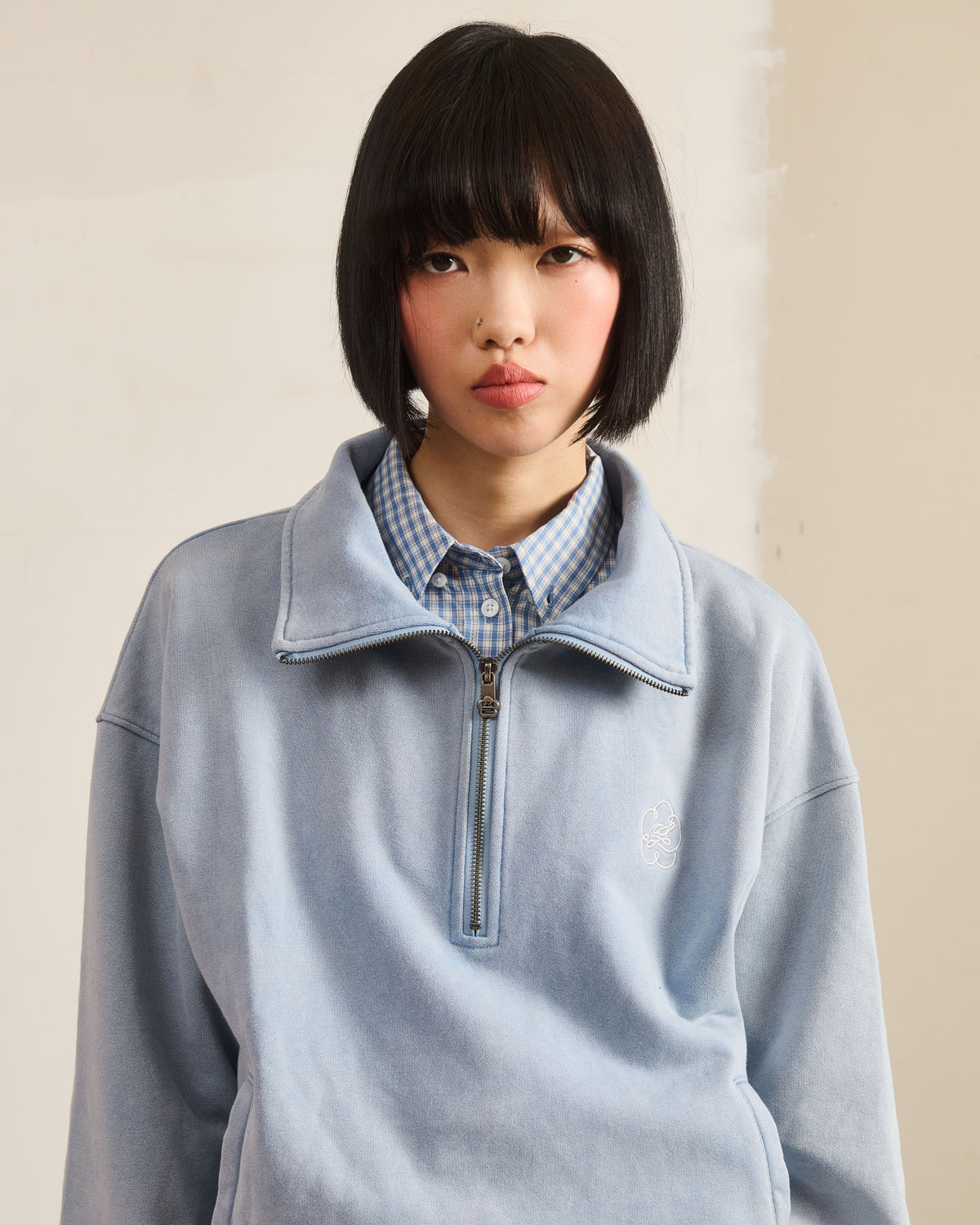 A person with a straight black bob wears the Sundae School SKY BLUE HESTER QUARTER ZIP over a blue and white gingham shirt, standing against a plain off-white background.