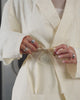 A person showing off the details of the IVORY LINEN GORUM BLAZER by Sundae School stands in the center of an empty subway car, hands behind their back and facing forward. The subway features silver walls and blue seats.