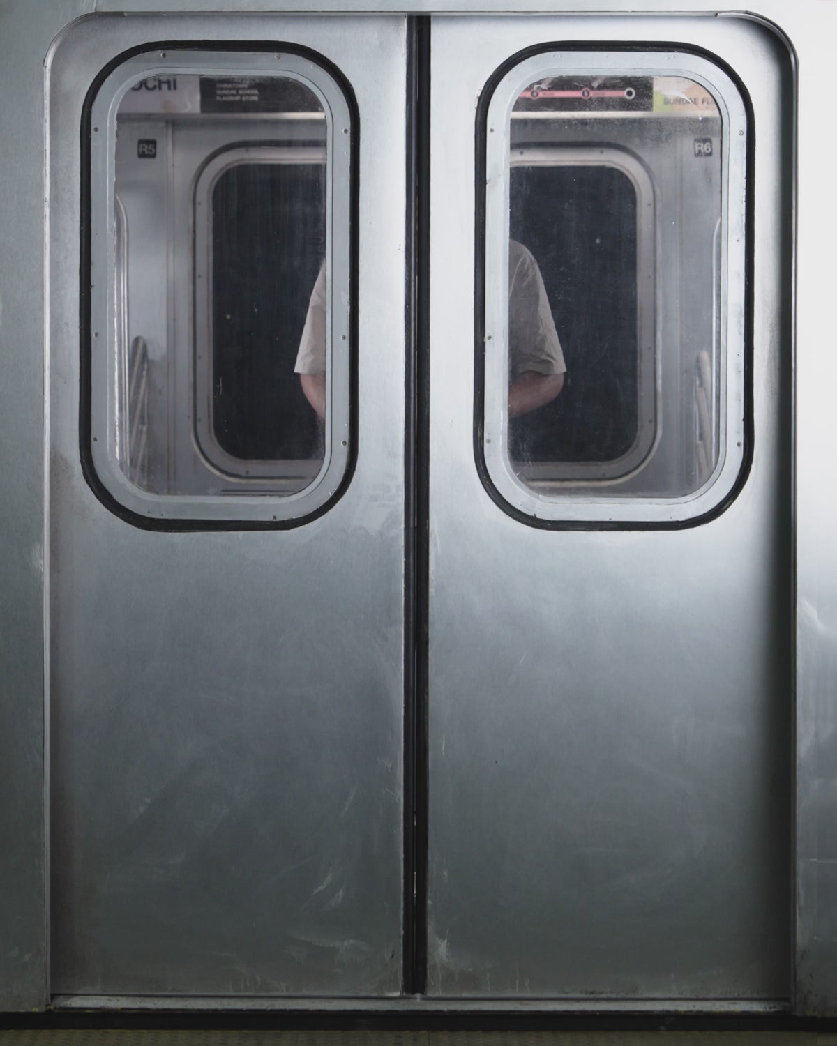 A person with curly hair stands in a subway car wearing Sundae School's WHEAT HANBOK LINEN SHIRT and WHEAT HANBOK LINEN PANTS, paired with a loose light beige short-sleeve suit and black shoes, hands in pockets, gazing slightly to the side.