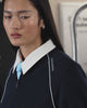 A woman with long dark hair and sunglasses wears the Sundae School NAVY OFFICE QUARTER ZIP, light blue tie, and dark shorts as she stands by subway doors beneath a map and a “117 Hester St” sign.
