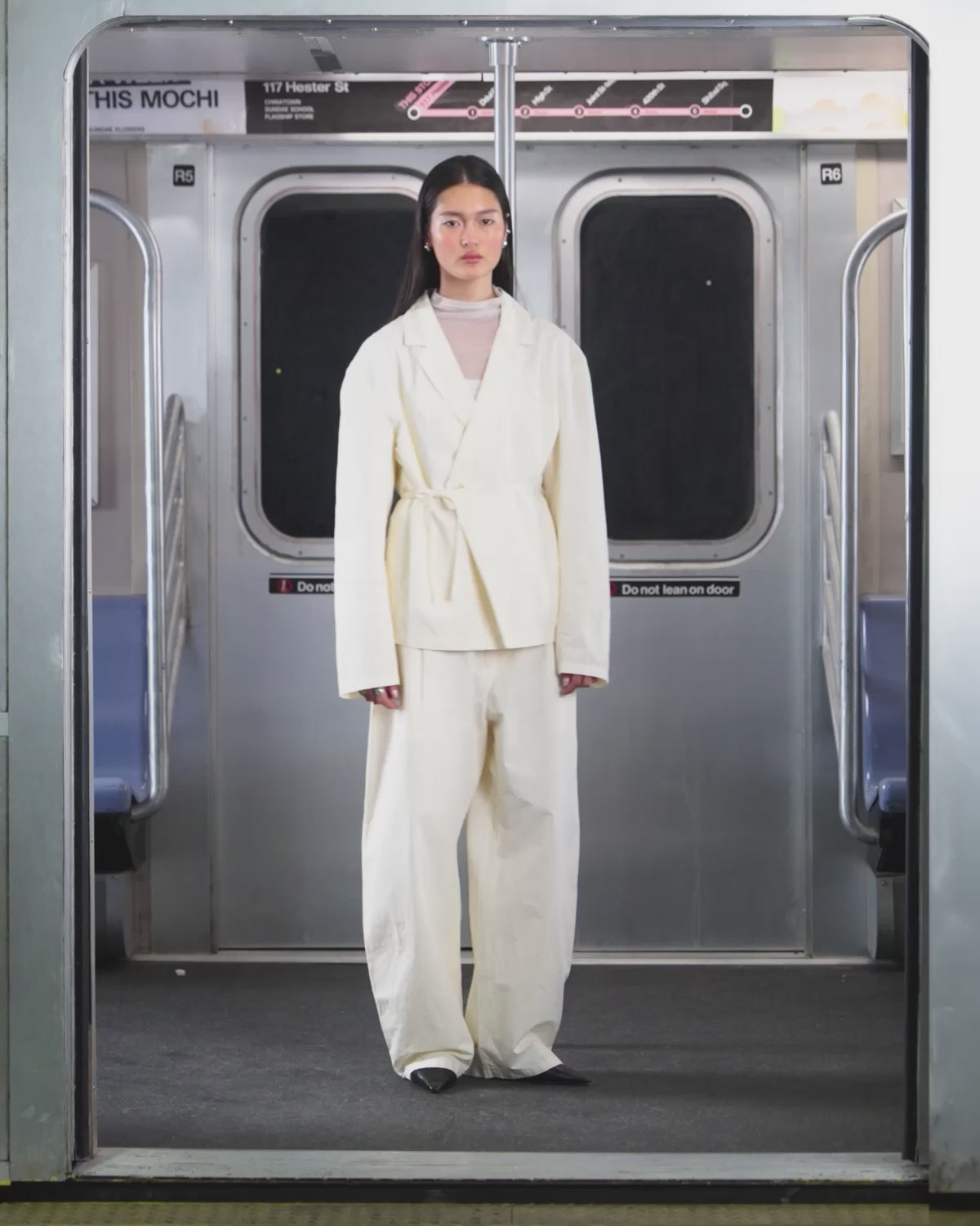 A person wearing the IVORY LINEN GORUM BLAZER and IVORY FLOWER NORIGAE LINEN TROUSERS by Sundae School stands in the center of an empty subway car, hands behind their back and facing forward. The subway features silver walls and blue seats.