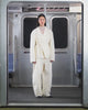A person wearing the IVORY LINEN GORUM BLAZER and IVORY FLOWER NORIGAE LINEN TROUSERS by Sundae School stands in the center of an empty subway car, hands behind their back and facing forward. The subway features silver walls and blue seats.