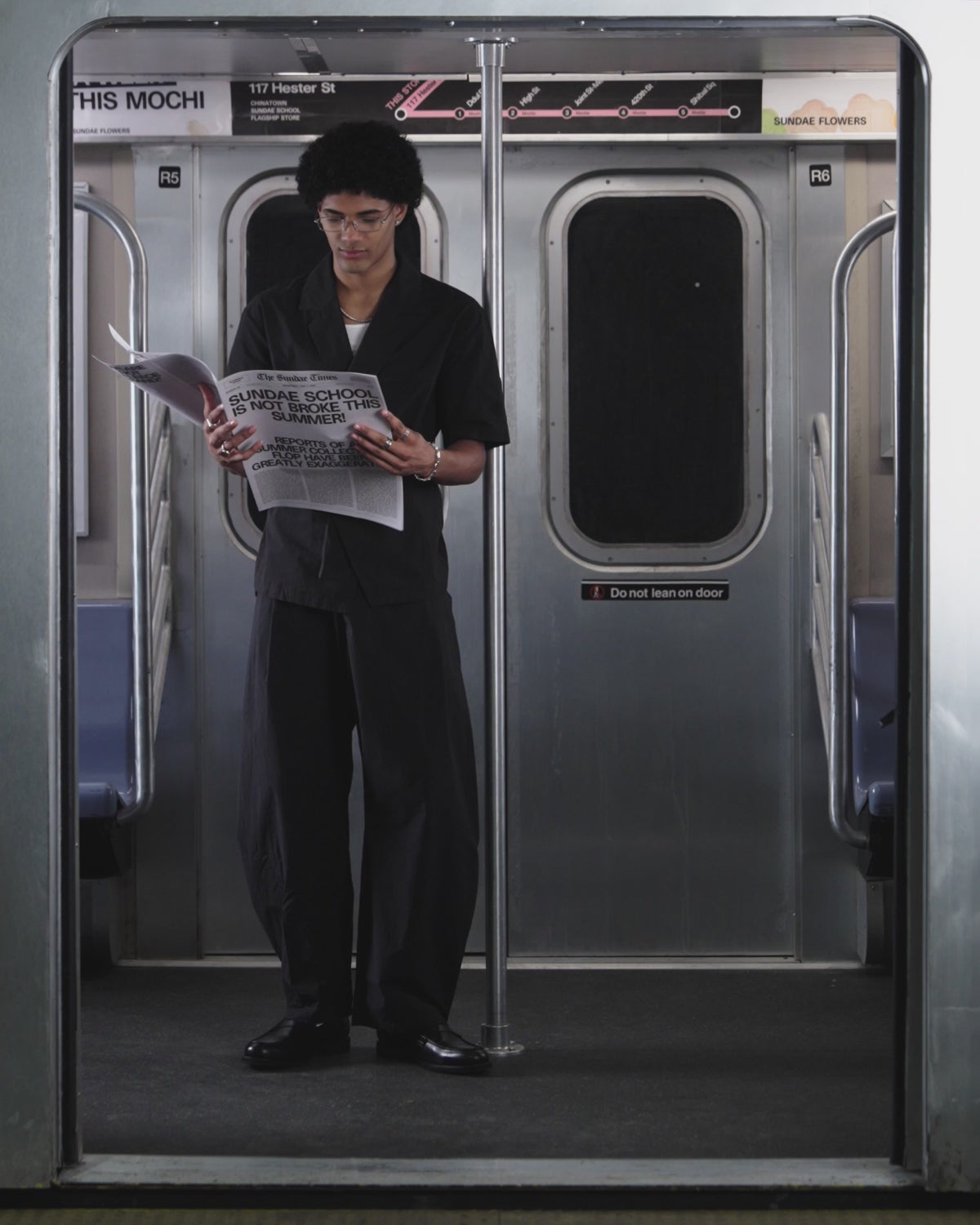 A young person with curly hair and glasses wears the BLACK HANBOK LINEN SHIRT by Sundae School in a subway car, standing beneath a 117 Hester St sign with a subway map beside it.