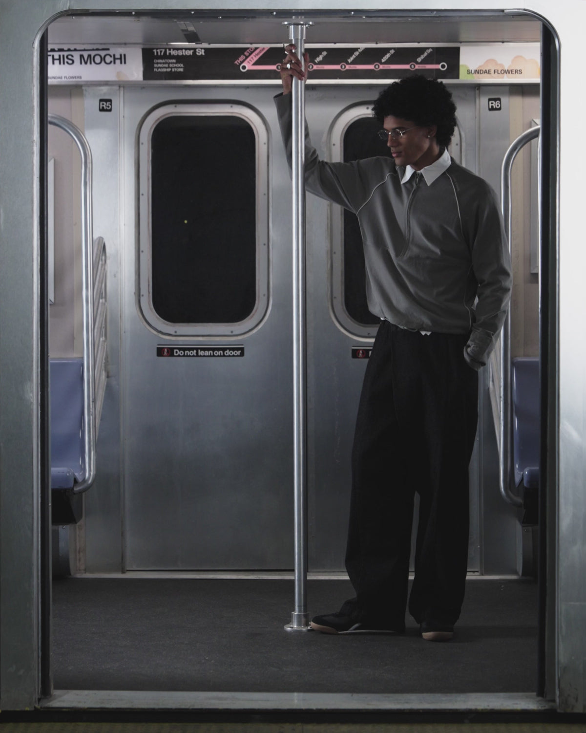 A person with curly hair, seen from behind in a Sundae School KHAKI OFFICE QUARTER ZIP, stands inside a subway train facing closed doors. Above the doors are a subway map and a sign that reads 117 Hester St.
