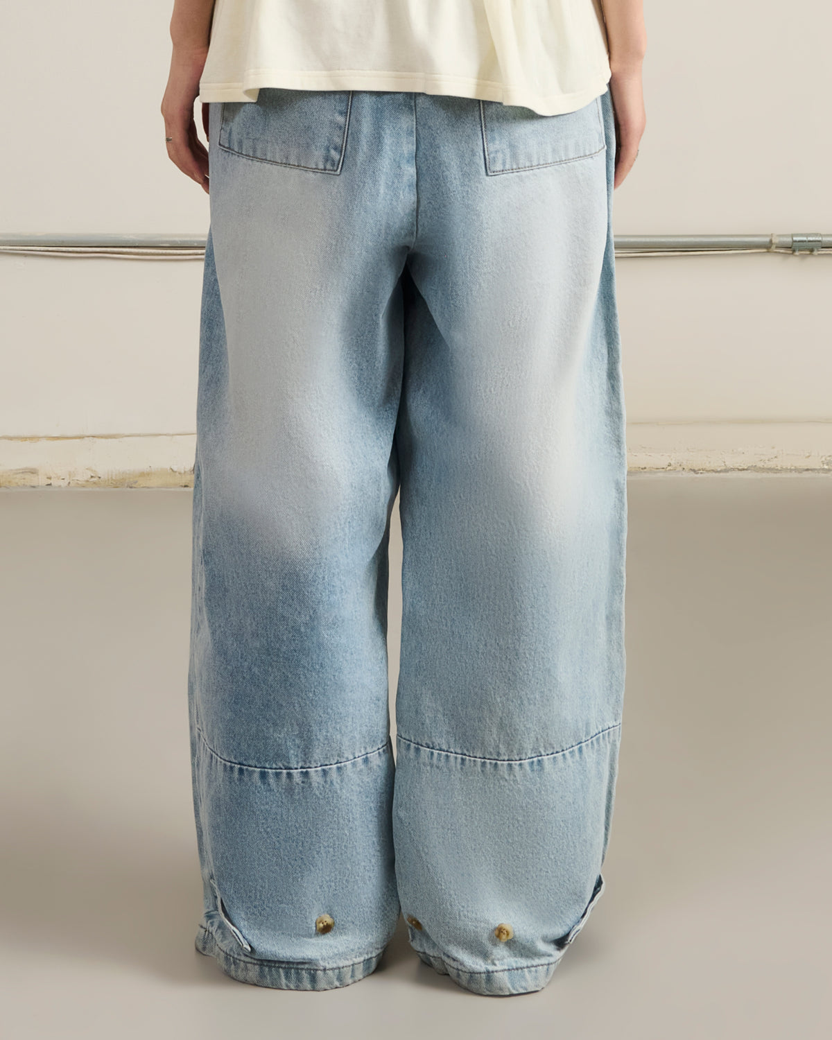 A person in Sundae School's SKY BLUE HANBOK DENIM PANTS with wide legs and large back pockets, shown from behind, stands on a smooth light-gray floor.