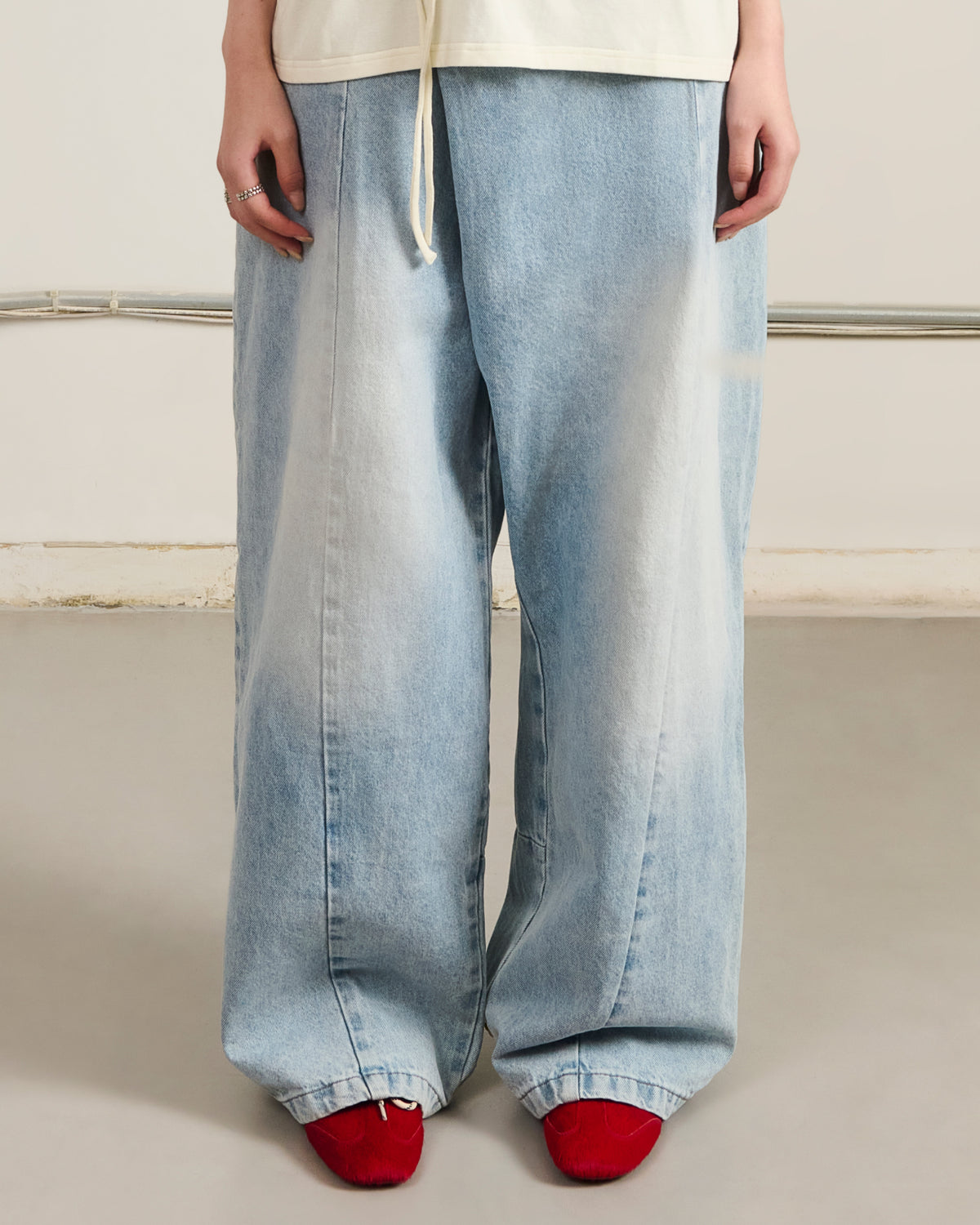A person stands indoors wearing Sundae School SKY BLUE HANBOK DENIM PANTS, loose-fitting with wide legs in a light wash, paired with bright red slippers. Only the lower half of their body is visible, hands resting by their sides.