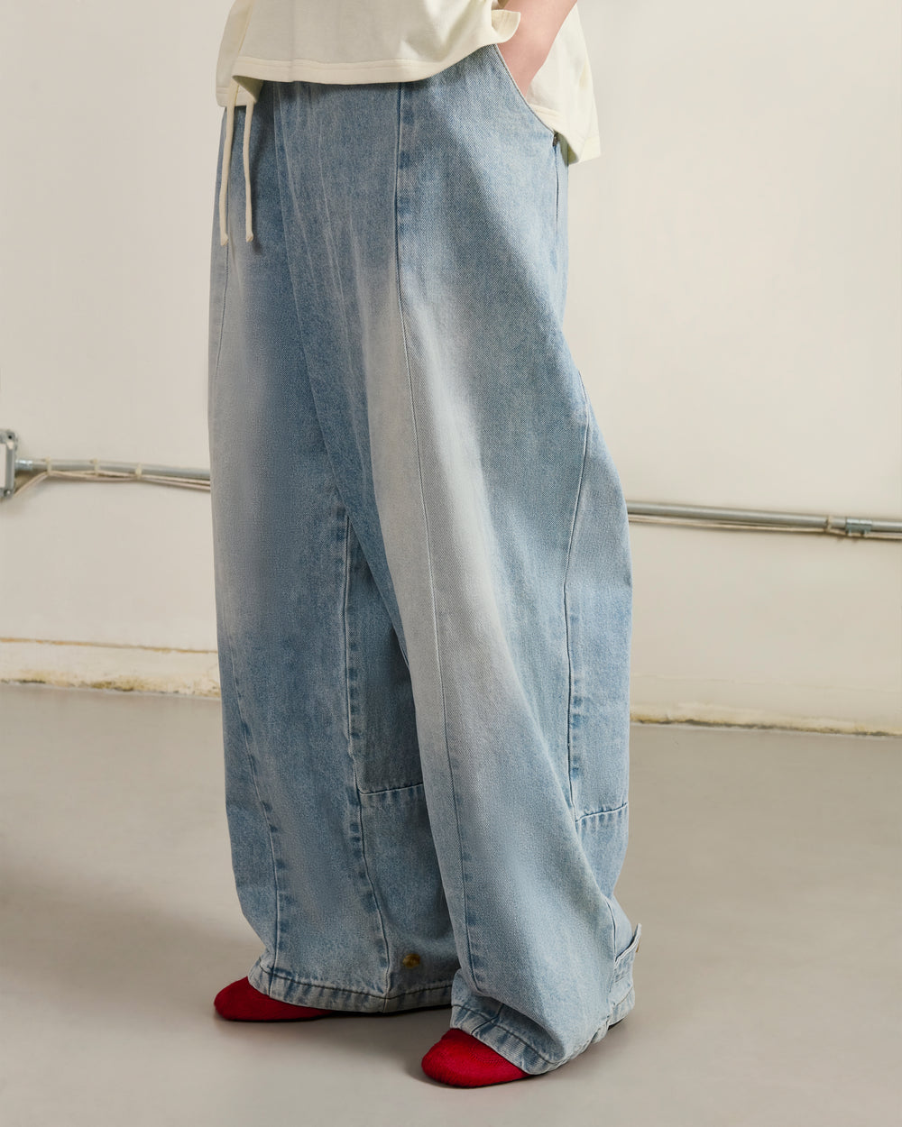 A person wears Sundae School’s SKY BLUE HANBOK DENIM PANTS with red socks, standing on a smooth, light floor. Their hands are in their pockets and a light shirt is partially visible on the upper body.